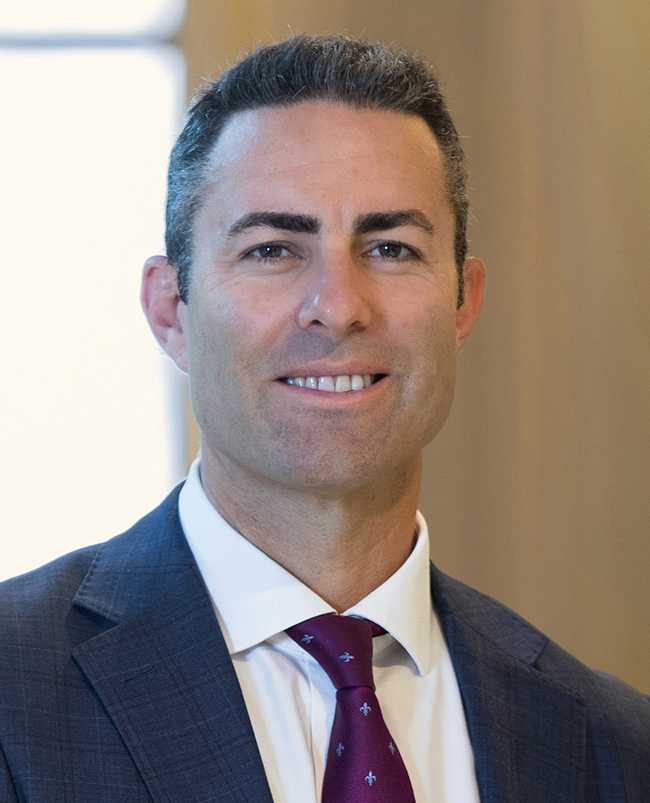 A headshot photograph of Brad, looking directly at the camera and smiling, wearing a dark suit, white shirt and light blue tie.