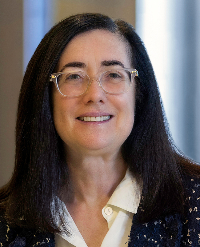 A headshot photograph of Gina, looking directly at the camera and smiling, wearing clear-framed glasses. 