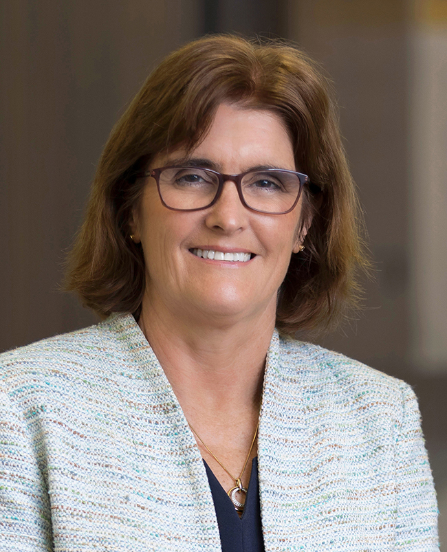 A headshot photograph of Michele, looking directly at the camera and smiling, wearing dark-framed glasses and a pale grey jacket.