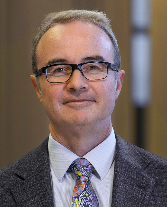 A headshot photograph of Scott, looking directly at the camera and smiling, wearing dark-framed glasses and a dark suit.